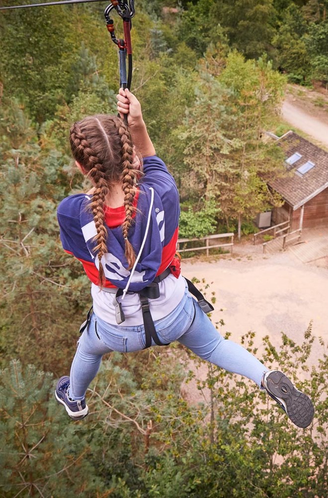 Teenage girl ziplines high above trees and wooden buildings below 