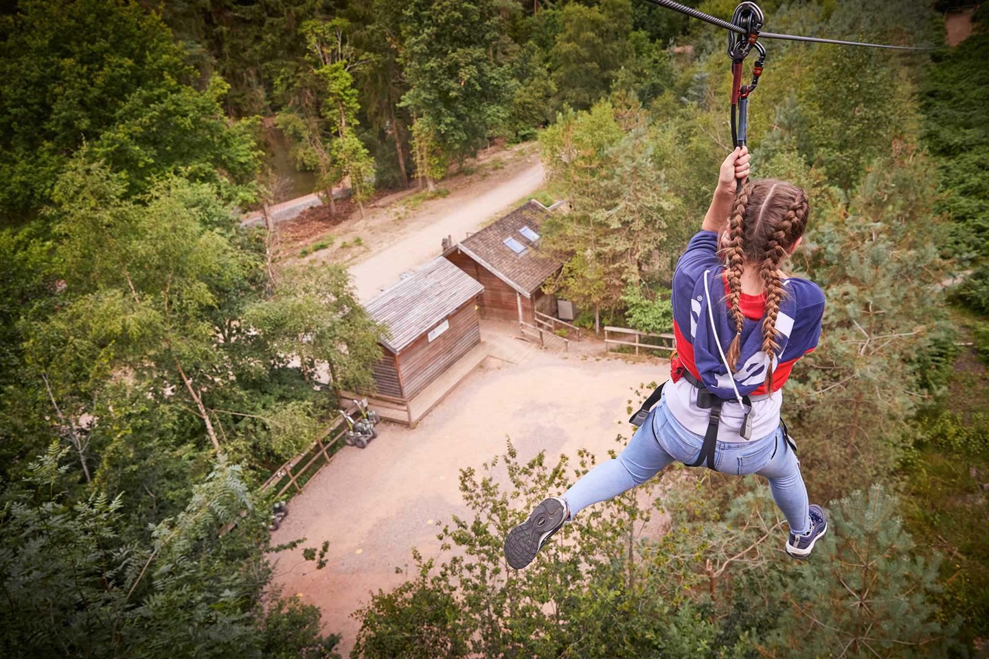 Teenage girl ziplines over trees and wooden buildings below 