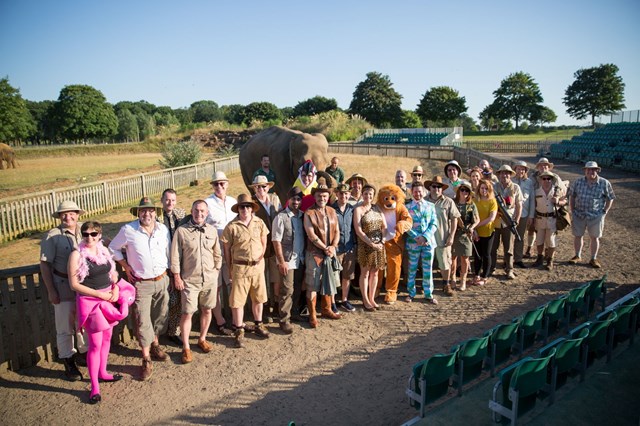 Large group in fancy dress stand in front of elephant and keeper in expansive grassy enclosure 